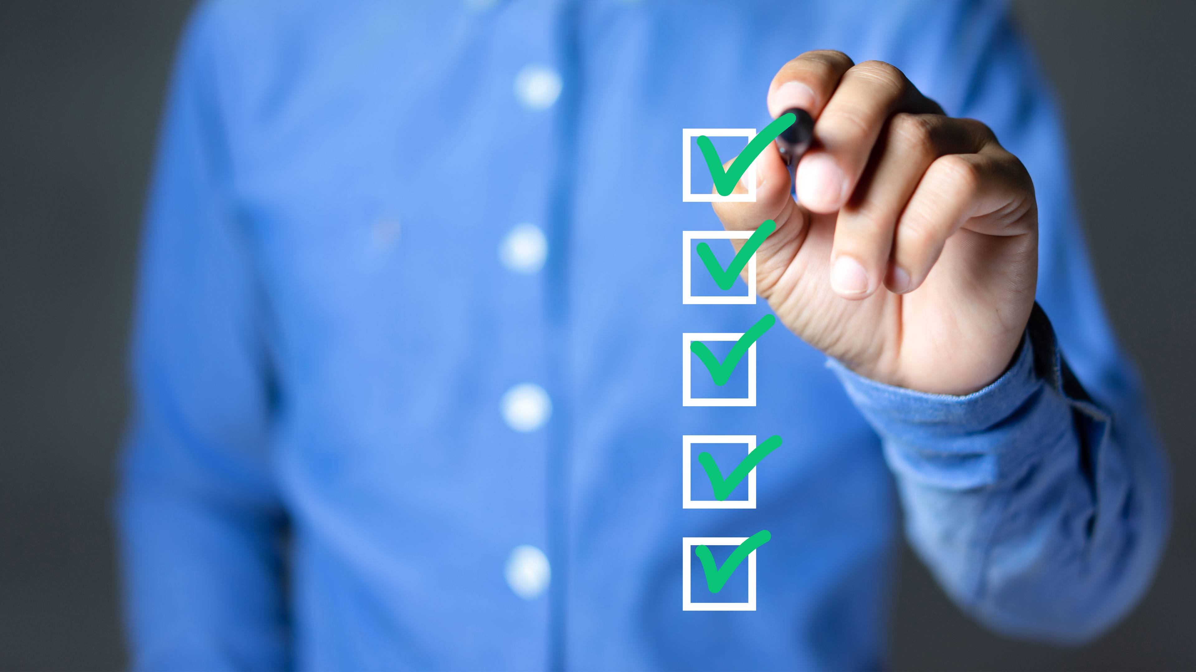 You can see the upper body of a man wearing a blue shirt and holding a pen in his hand. He uses it to tick off white squares, which are placed over the picture in a composition. All the squares are ticked in green.